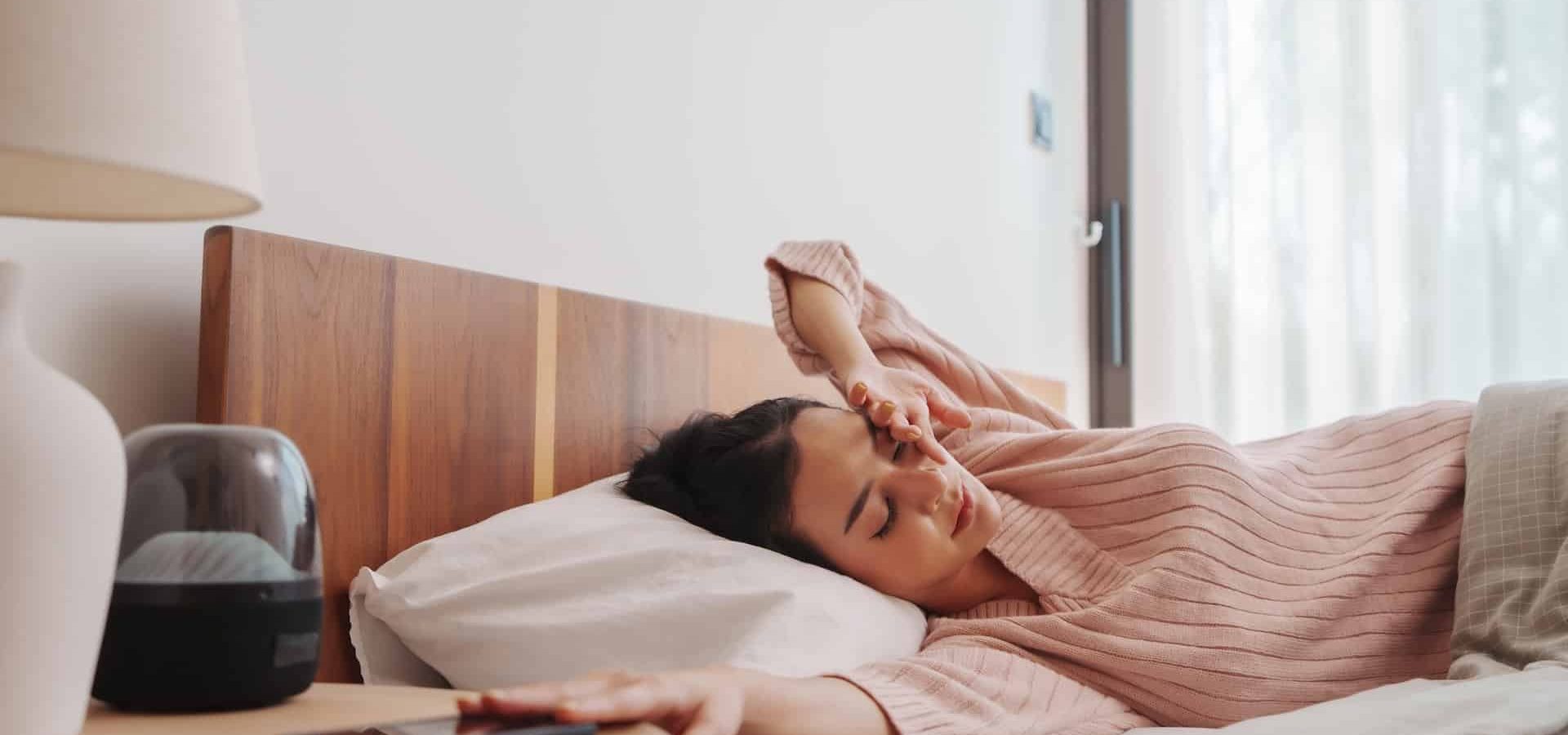 Young Asian woman in a soft pink sweater lying in bed with a headache, hand on forehead, with a serene expression and natural light pouring in from the window.