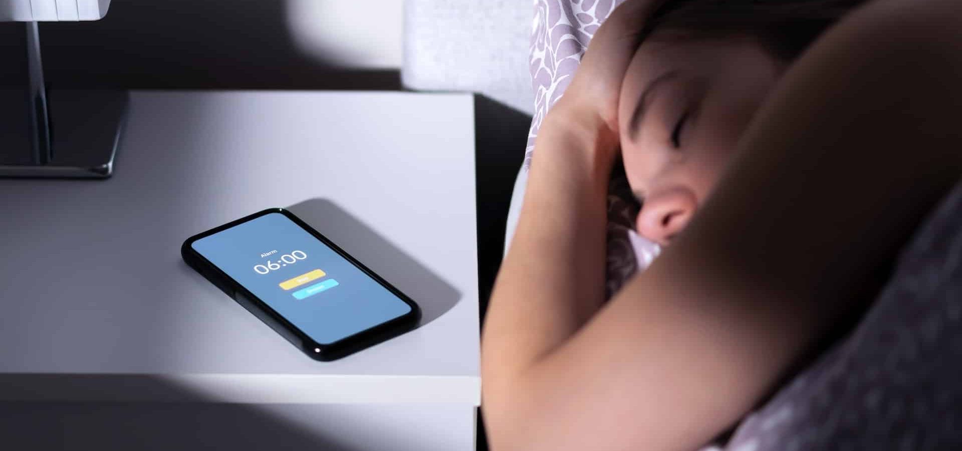 Phone alarm waking up tired sleeping woman in bed at night or morning. Cellphone on table with clock timer and snooze button. Oversleeping person, late from work. Sleepy, lazy or overslept lady.